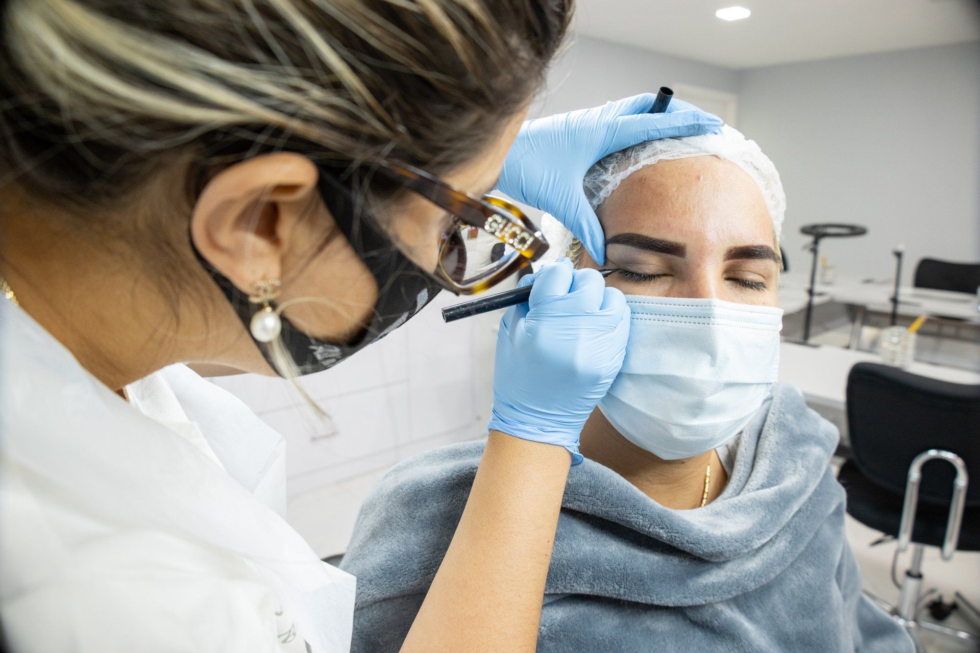 Woman getting eyebrow procedure by professional in a clinic, both wearing masks and gloves.