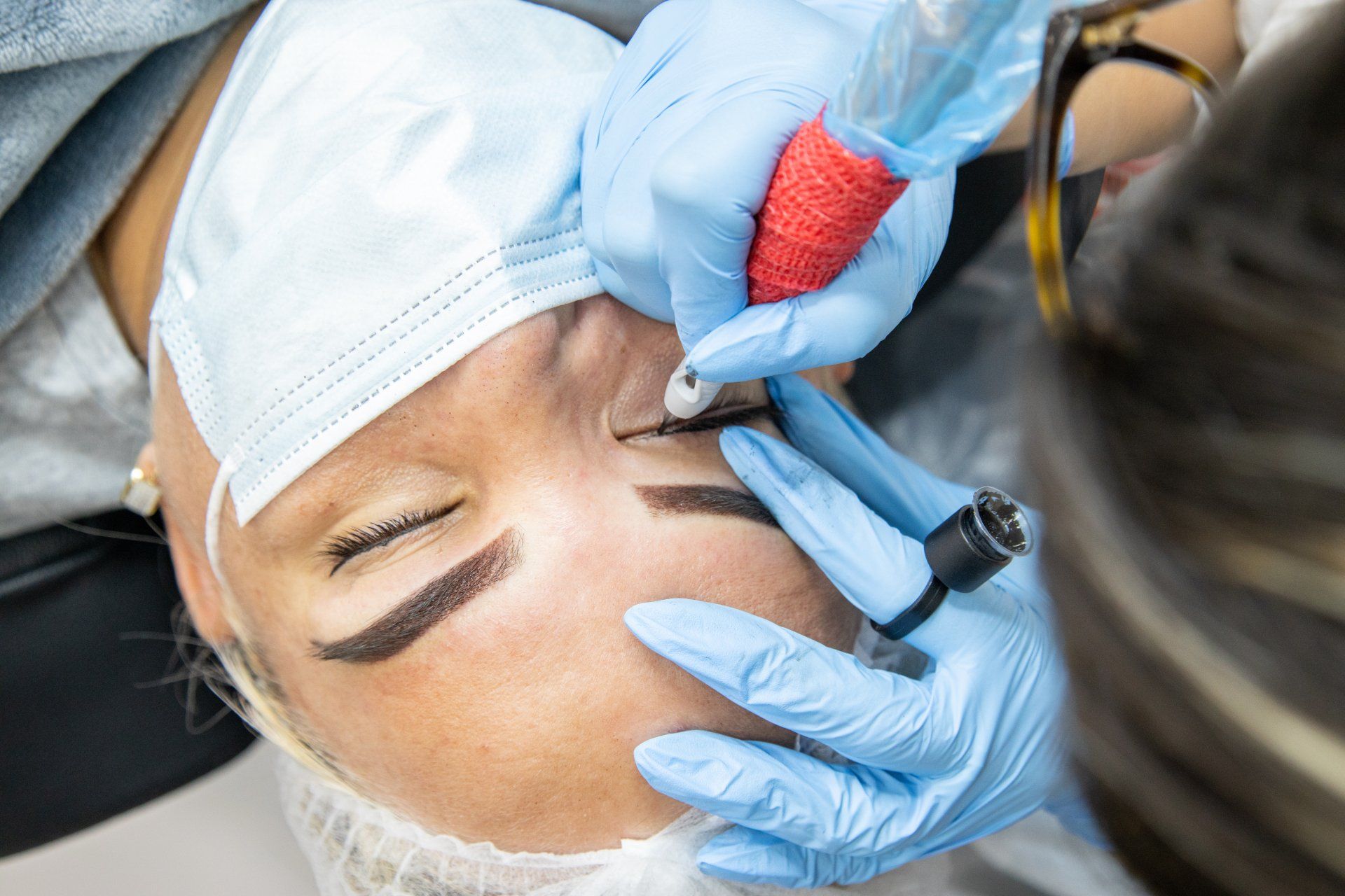 Person getting eyebrow tattoo; artist in blue gloves, mask, and red tattoo gun.