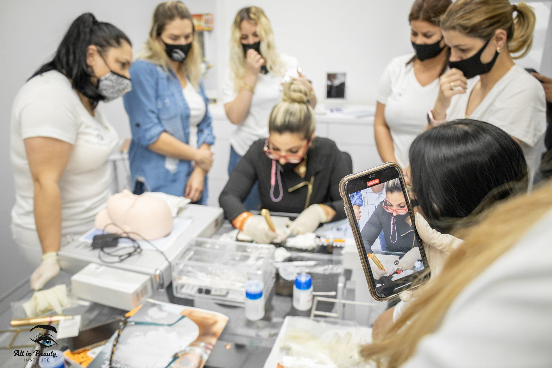 Group of women in masks watching a demonstration, one filming on a phone, in a brightly lit room.