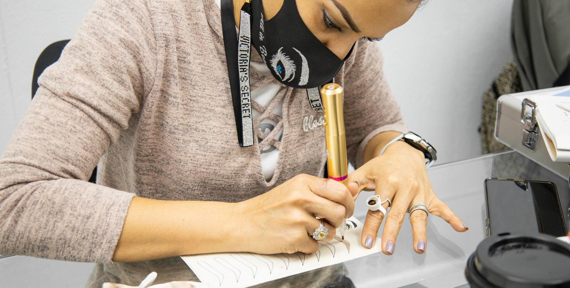 Woman wearing a mask, writing with a gold pen, rings on her fingers, indoors.