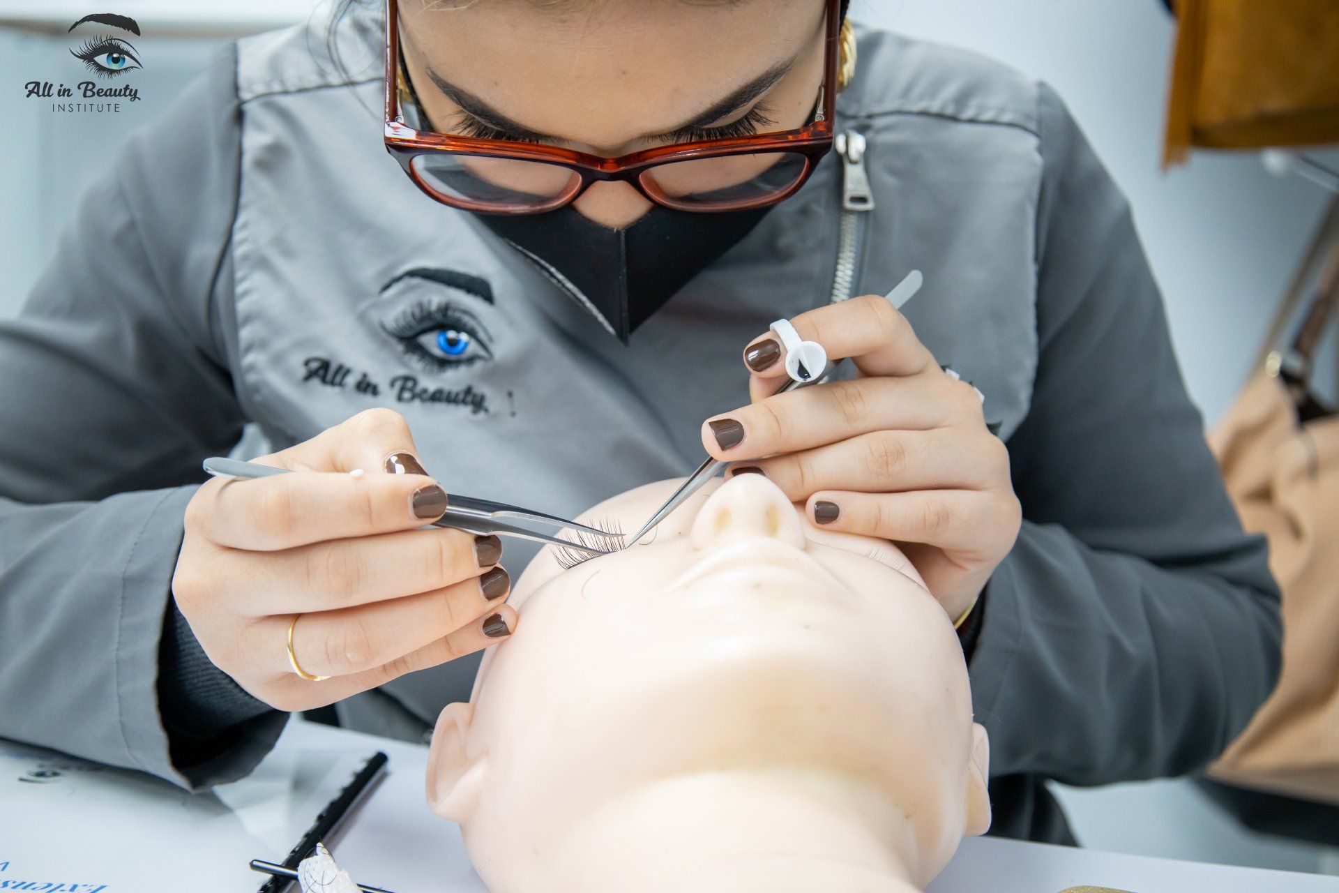 A woman with glasses and a mask applying eyelash extensions on a mannequin.