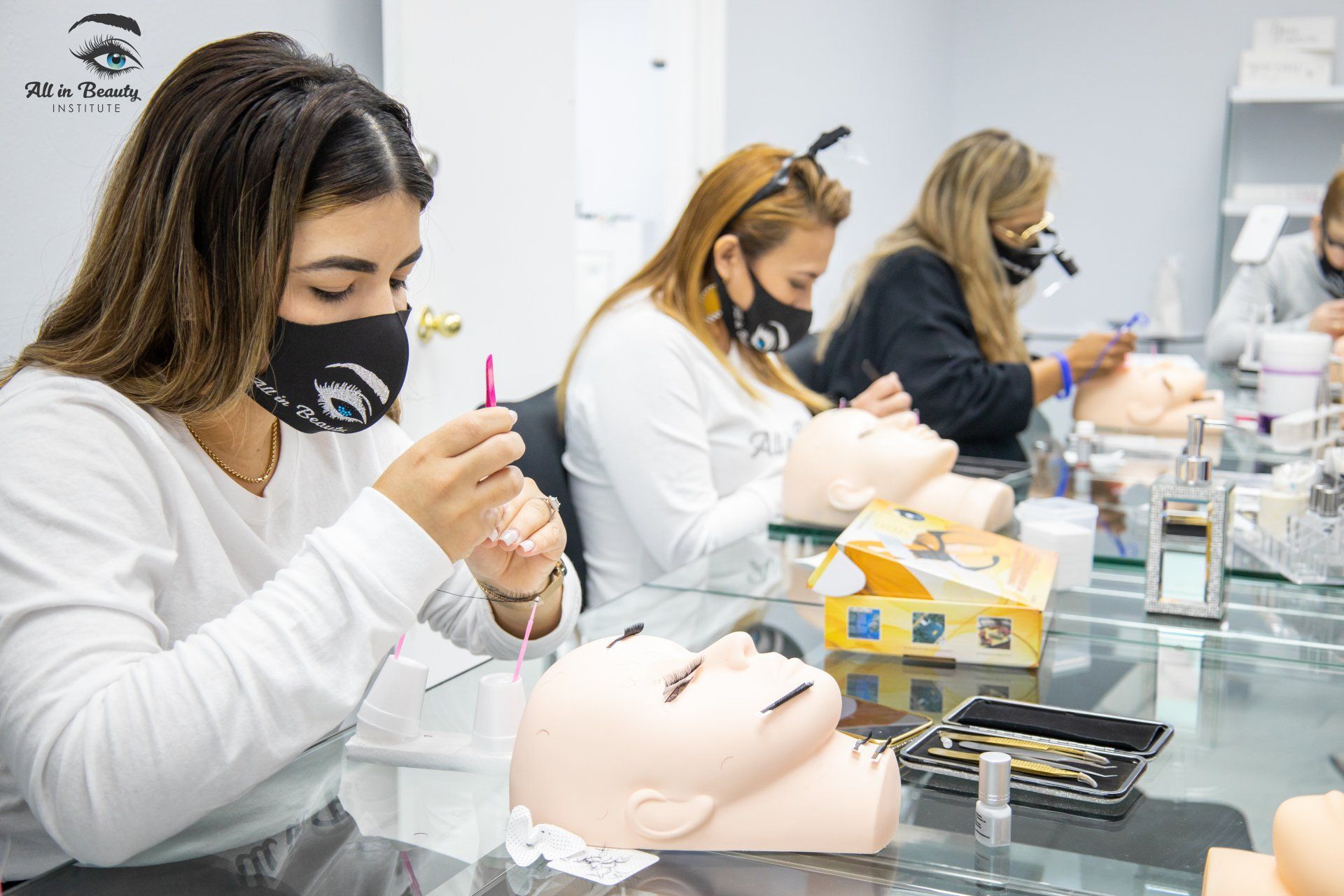 Students in masks practice applying eyelashes on mannequin heads at a class.