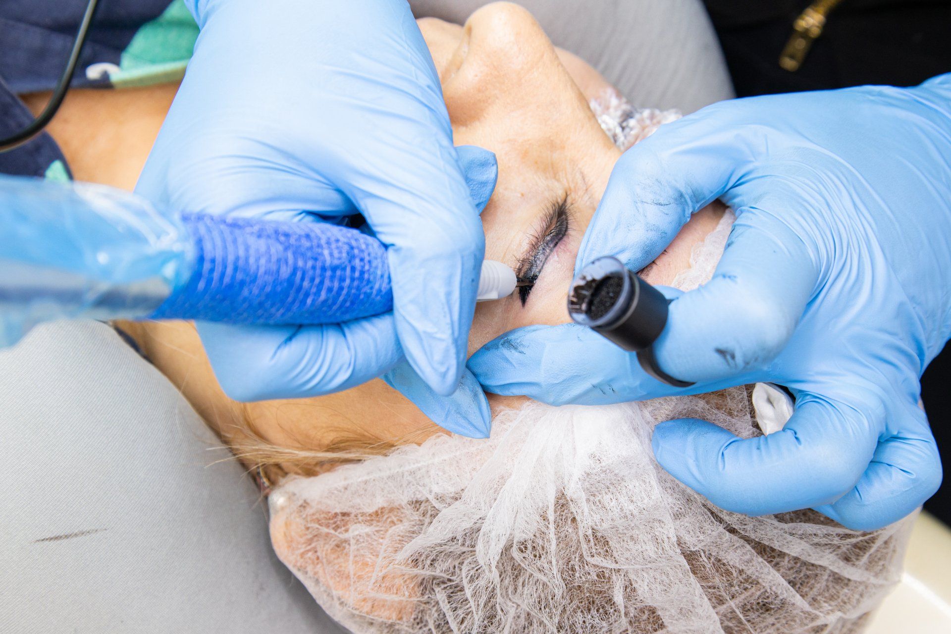 Hands in blue gloves performing eyebrow microblading on a woman's face.