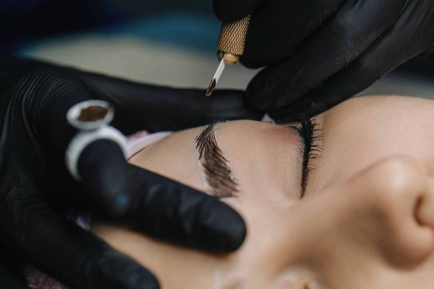 Person receiving eyebrow microblading; technician's gloved hands, dark ink, close-up.
