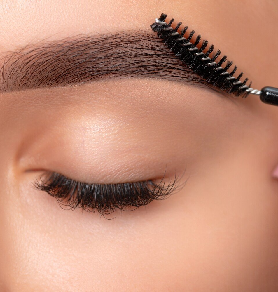 A person with closed eyes, brushing their filled-in eyebrow with a spoolie.
