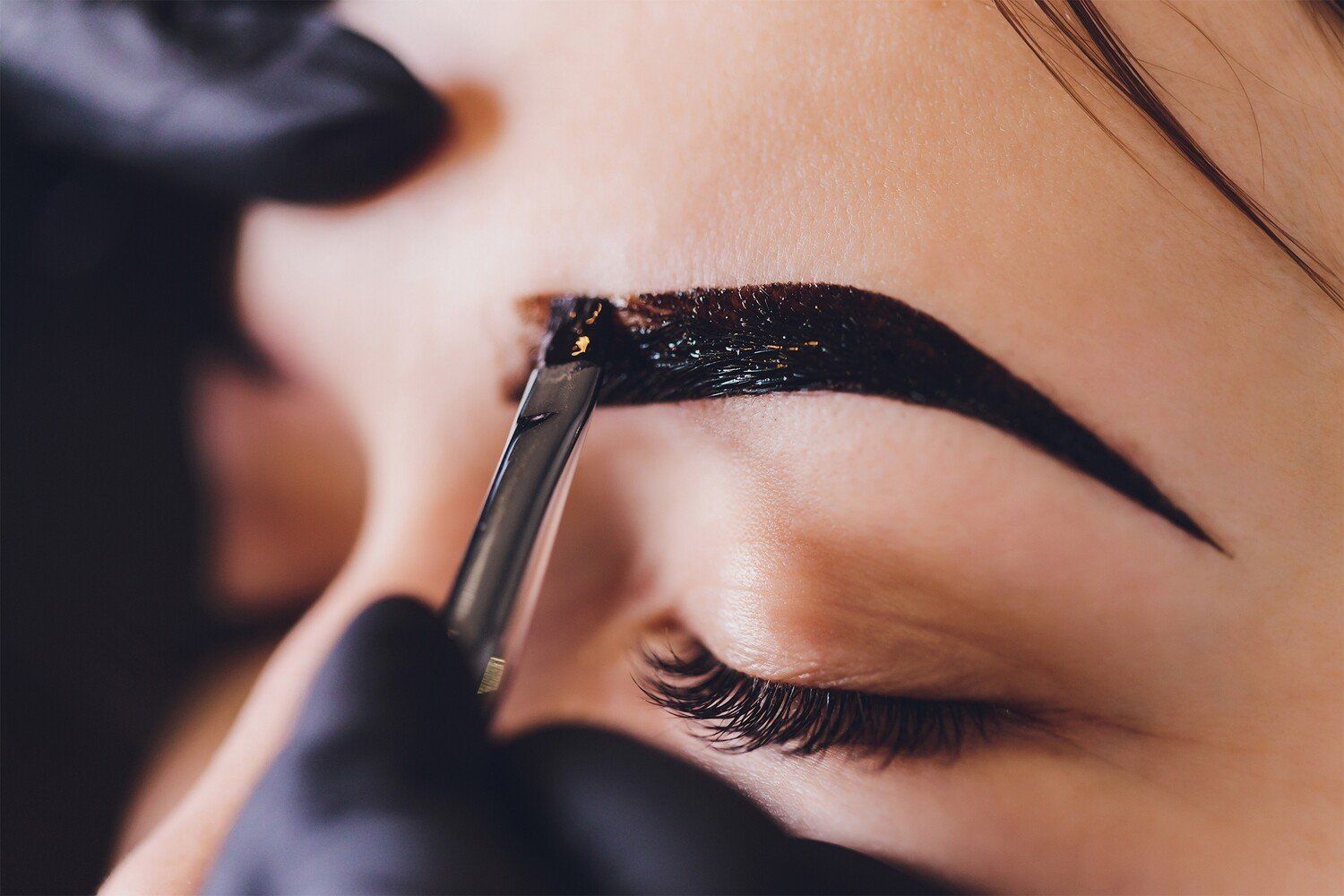 Person having dark eyebrow dye applied with a spatula by gloved hand.