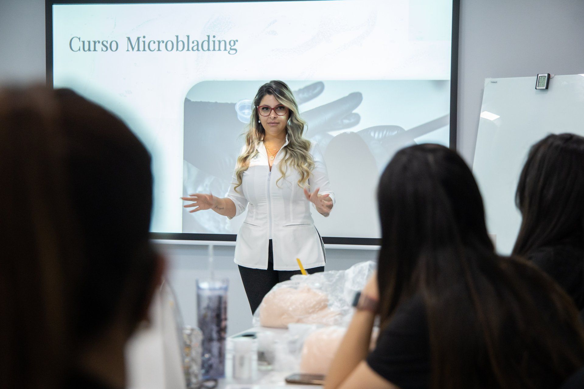 Woman in white coat teaching a microblading class. Students listen in a classroom.
