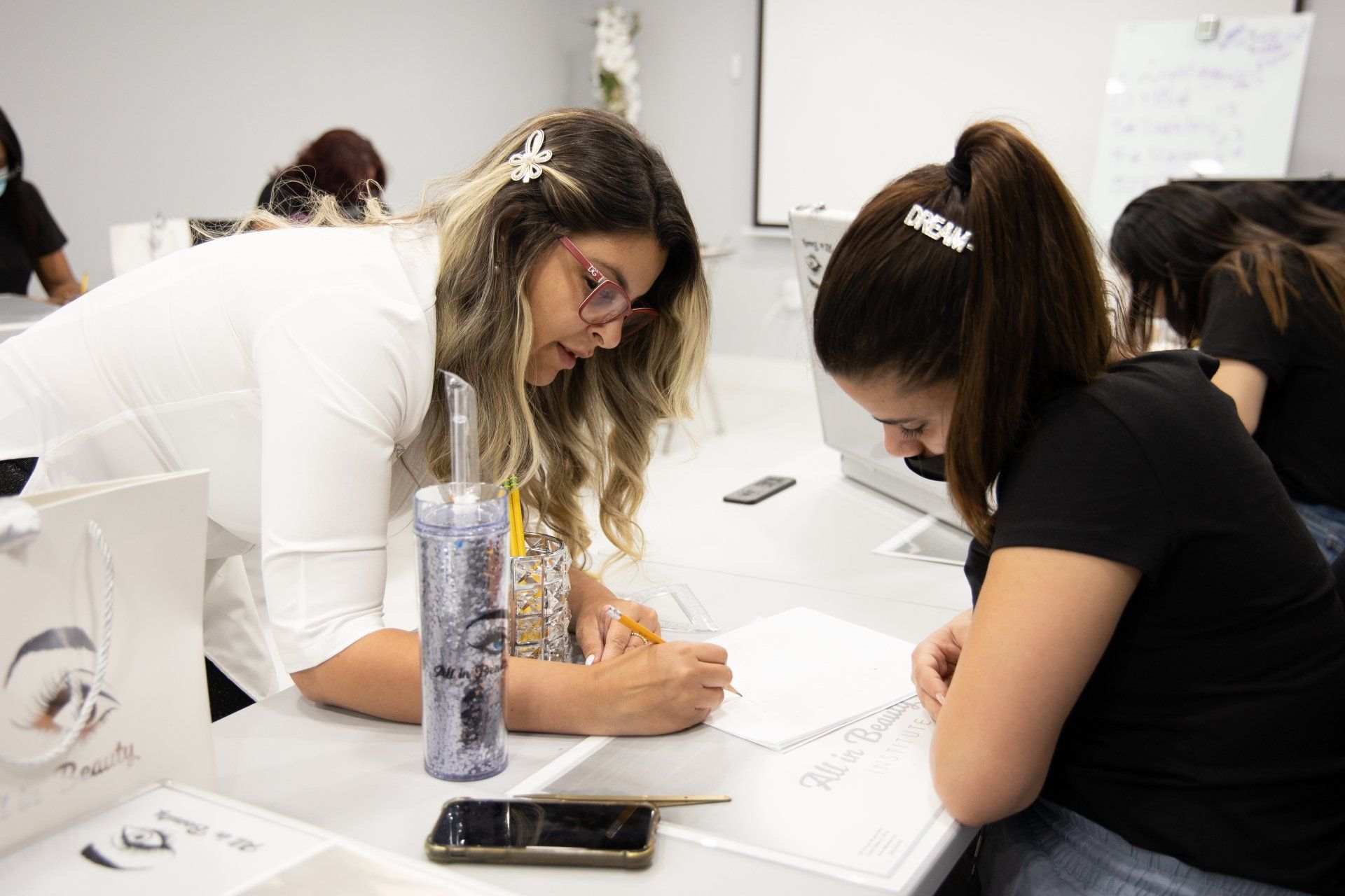 Two women at a table drawing, one helping the other. Classroom setting.
