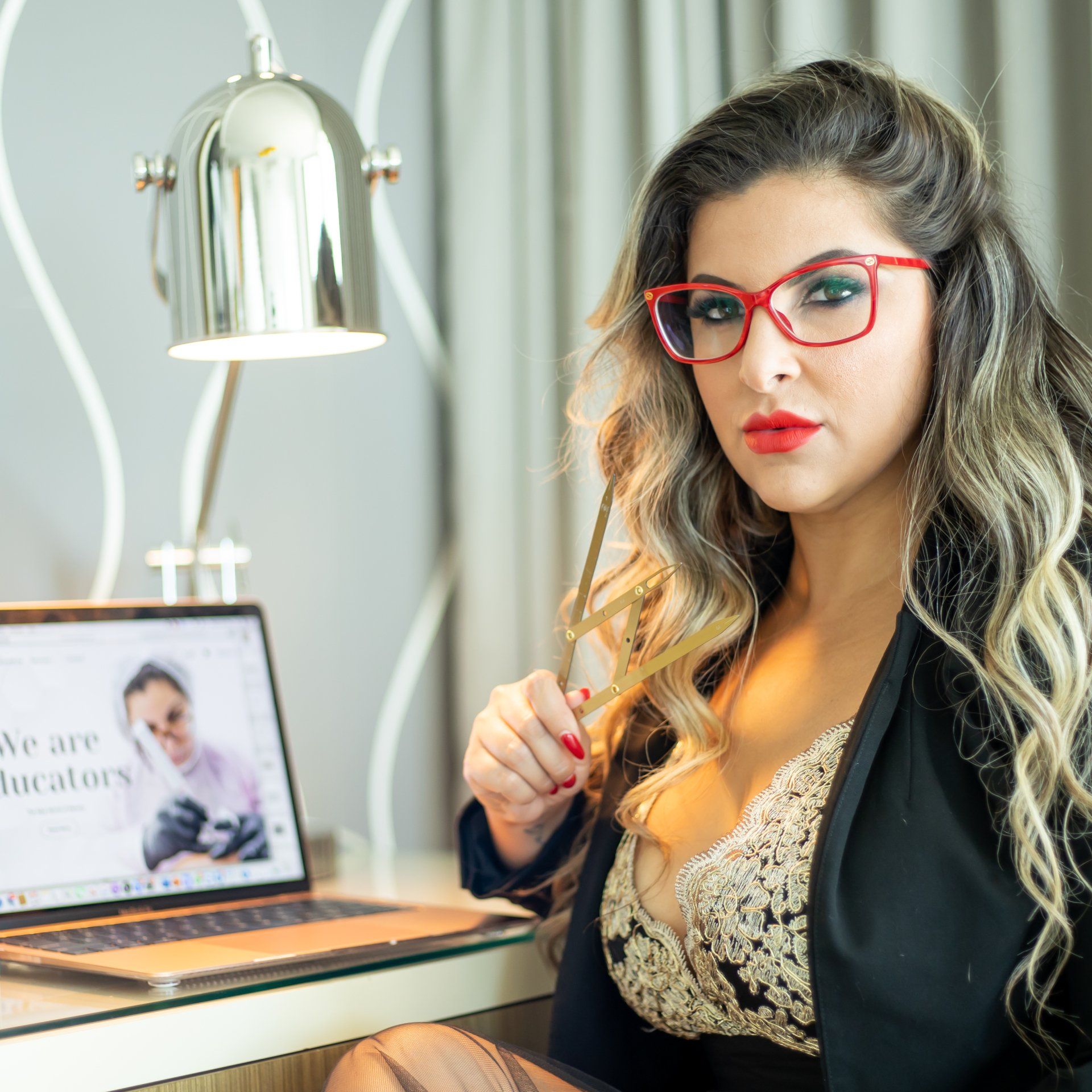 Woman in red glasses at a desk with a laptop, wearing a blazer, looking at the camera.