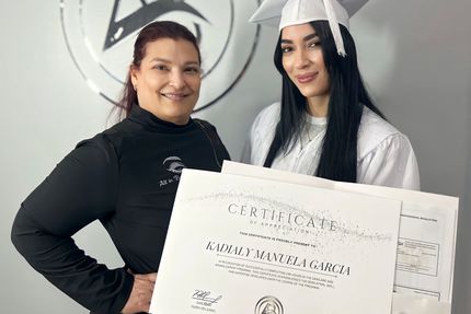 Woman and graduate holding diplomas, smiling. White graduation gown, cap. Gray background, logo.
