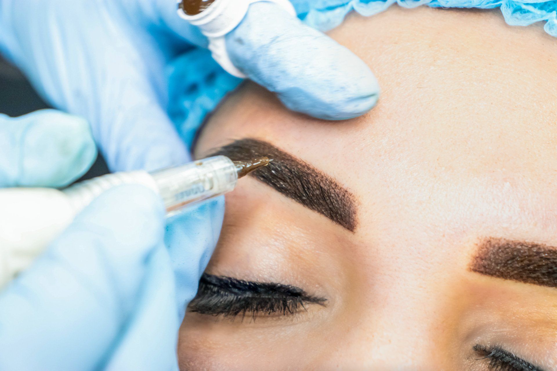 Eyebrow tattoo procedure in progress; gloved hands, dark brown pigment, close-up.