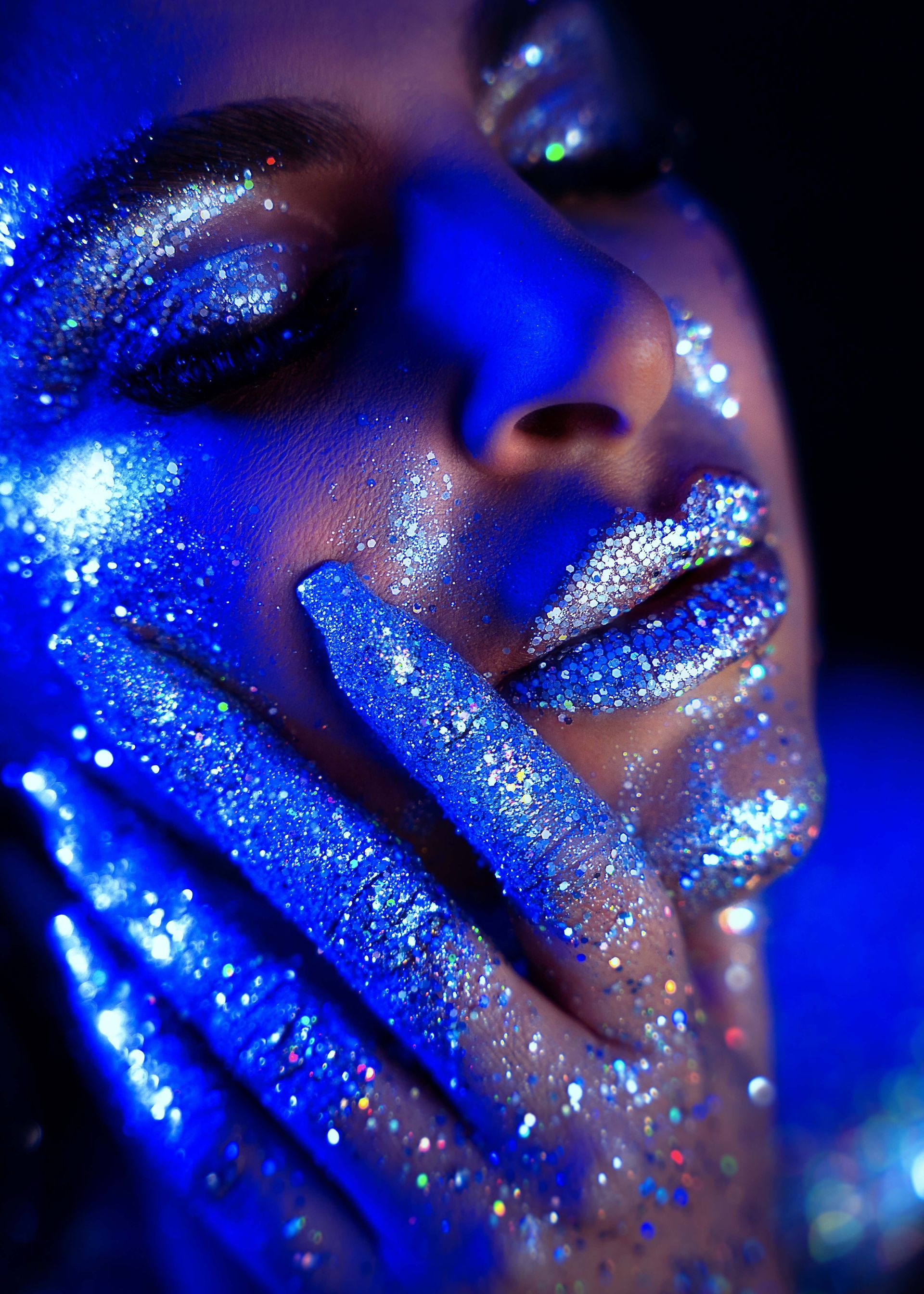 Woman with blue-toned skin and glitter on face and hand, lips are glittery, gazing downward.