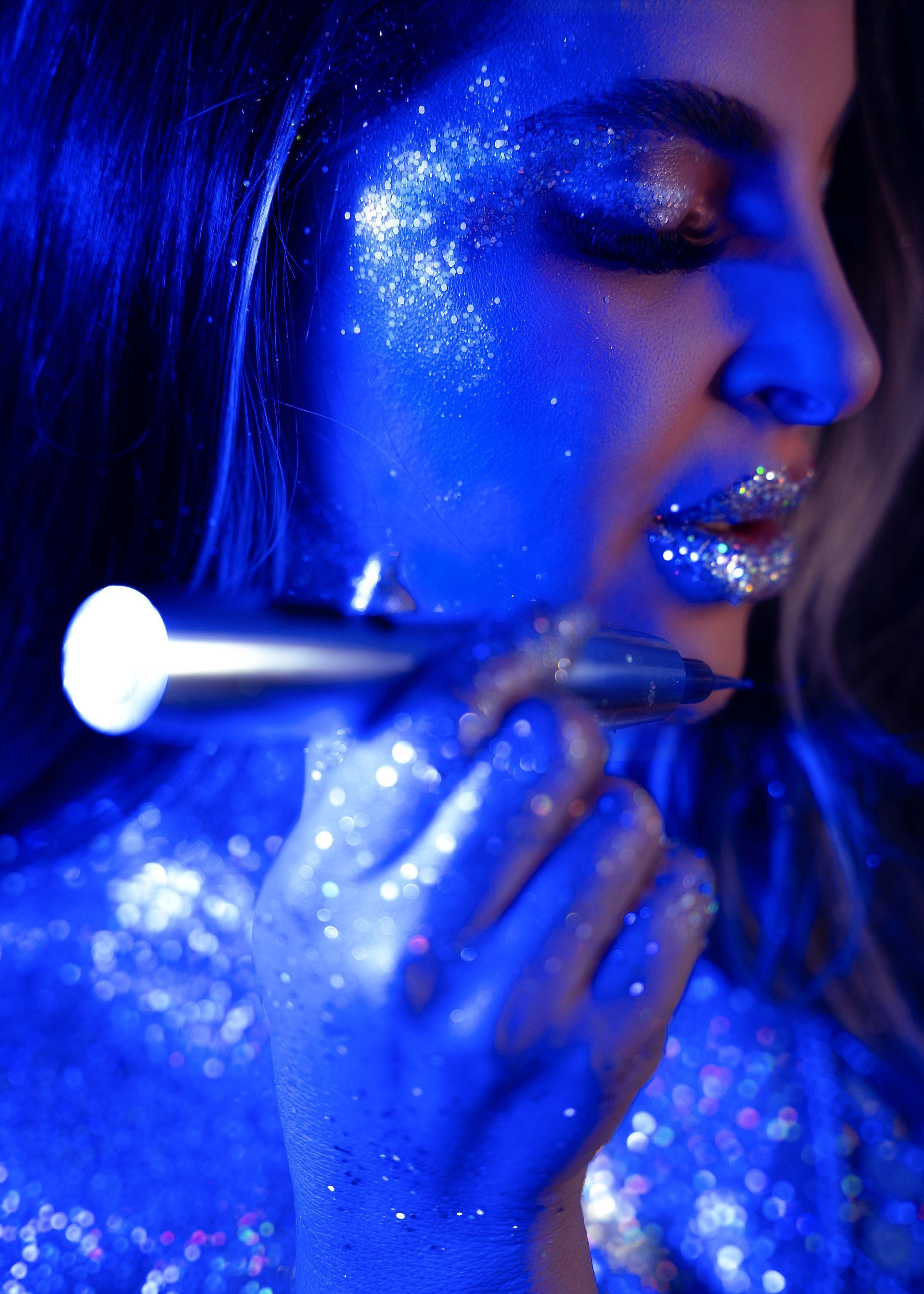 Woman with glitter makeup, holding a lit wand, illuminated in blue light.