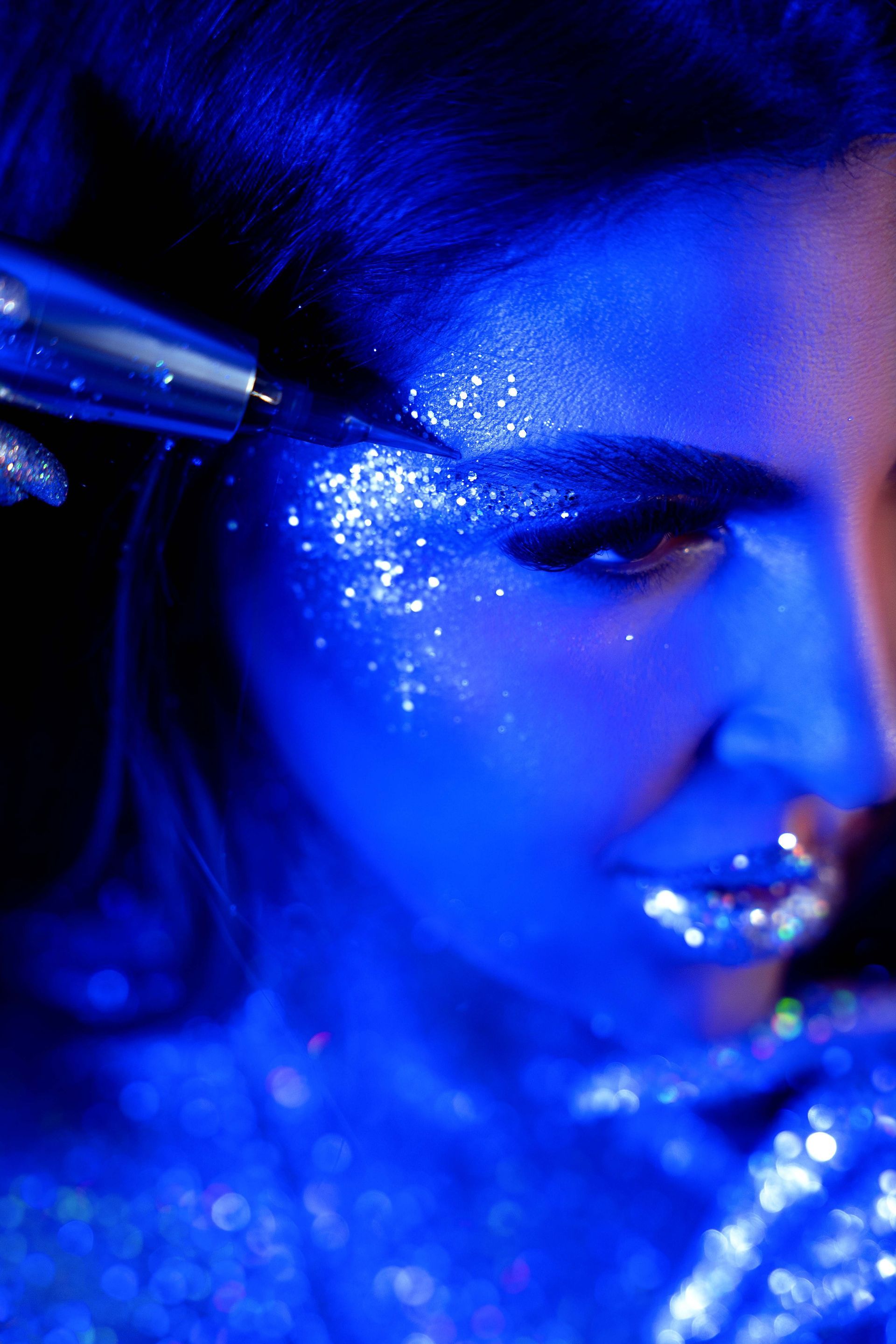 Woman with glitter makeup, applying eyeliner under blue light.