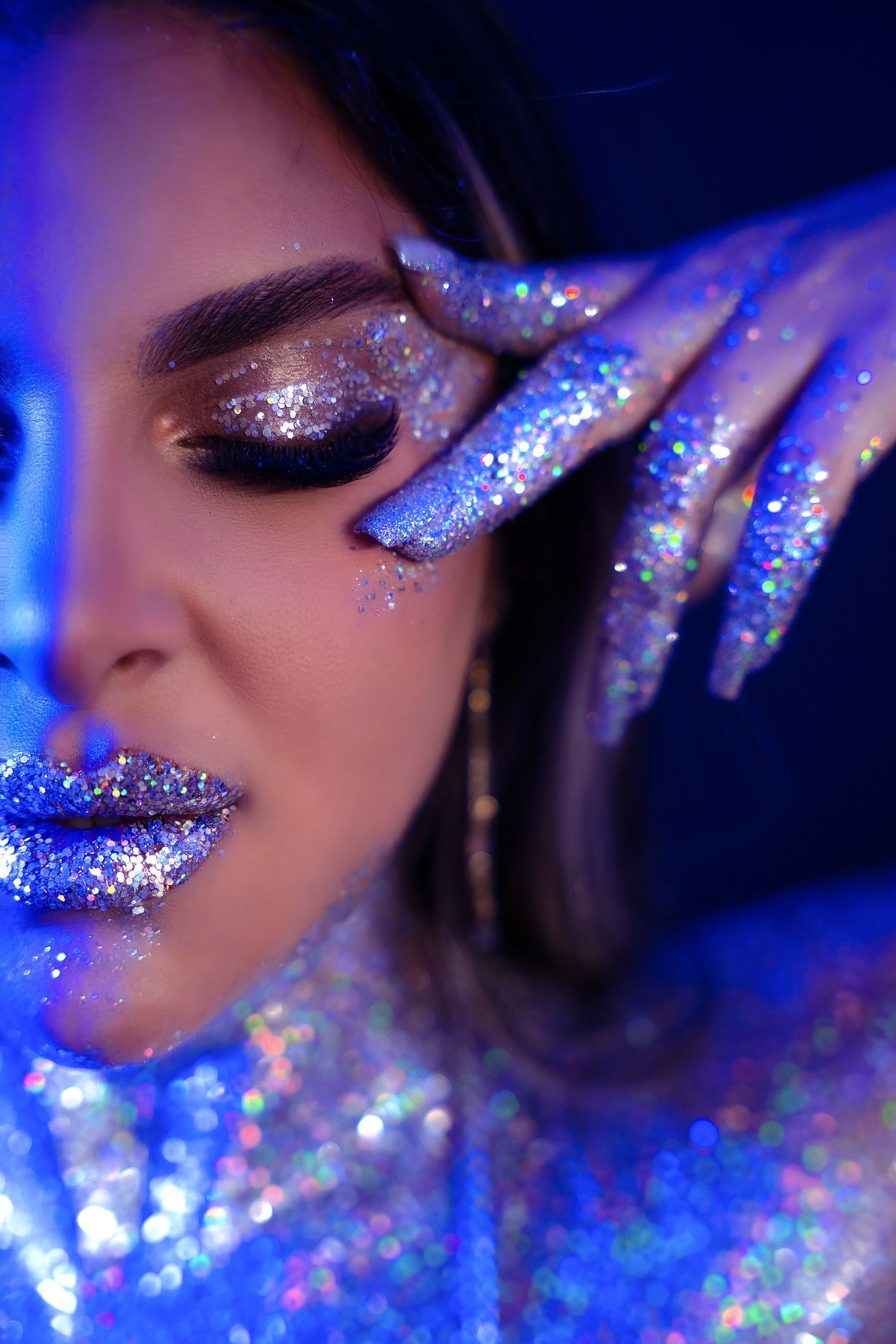 Woman with silver glitter makeup and hand to face, lit with blue light.