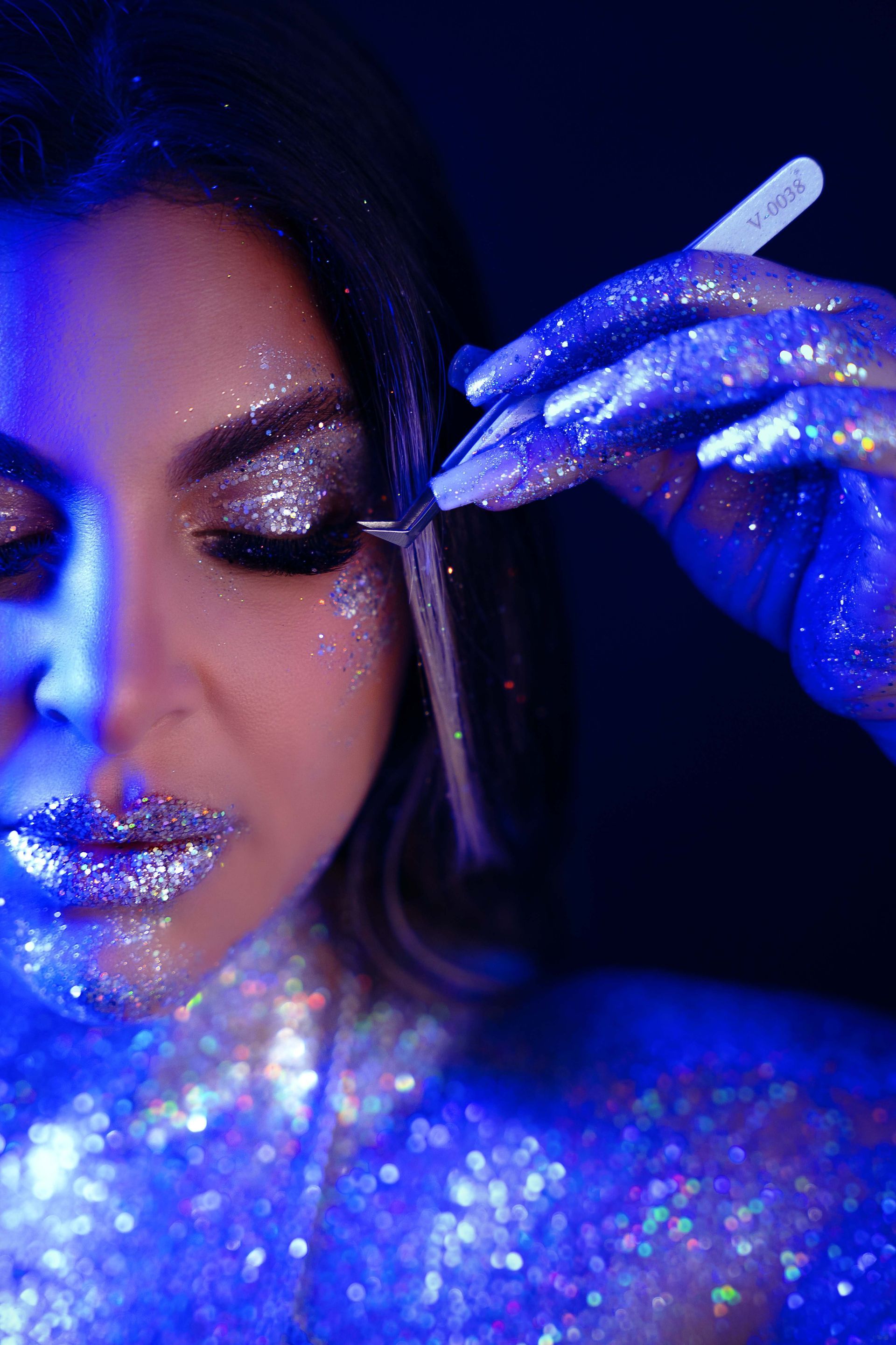 Woman with glitter makeup under blue light, applying false eyelashes.