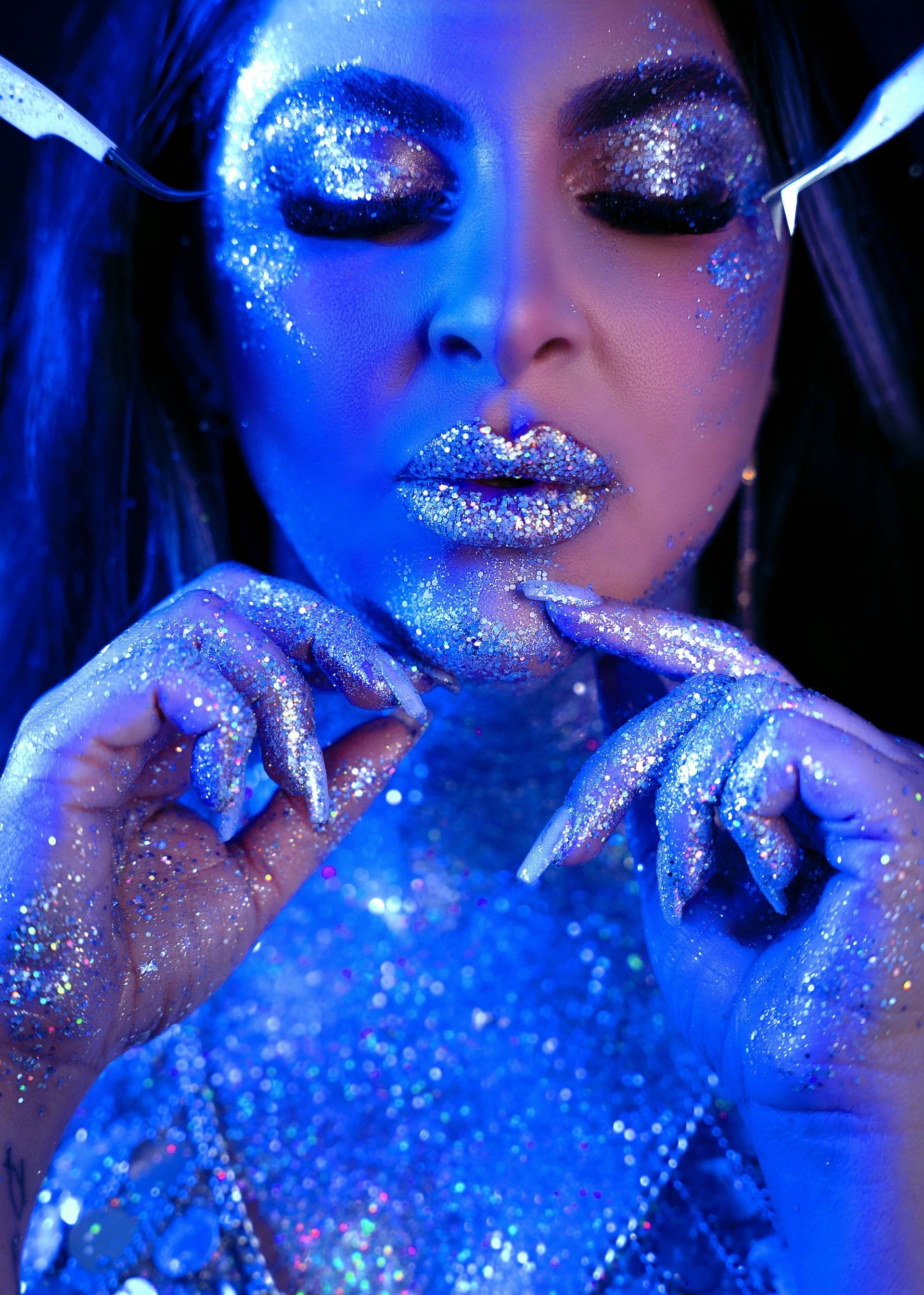 Woman with glitter makeup, lit by blue and purple light, fingers at lips.