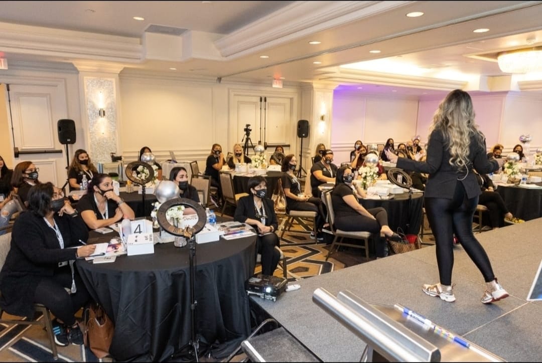 Woman speaking to a masked audience at a conference. Tables with lights, in a large room.