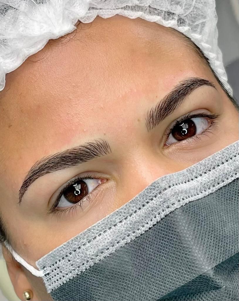 Close-up of woman's face; newly microbladed dark brown eyebrows; wearing surgical mask and cap; brown eyes.