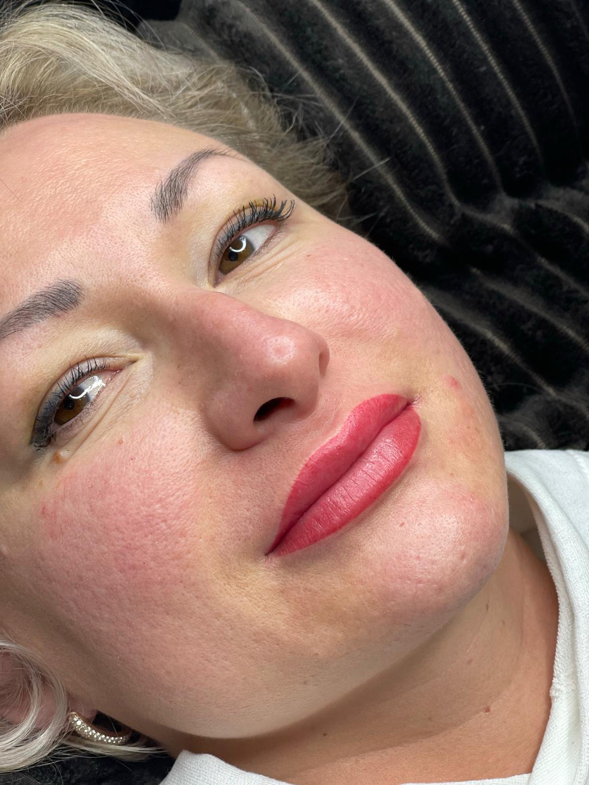 Woman with pink lip tattoo, light skin, and slight redness on cheeks, smiling softly.