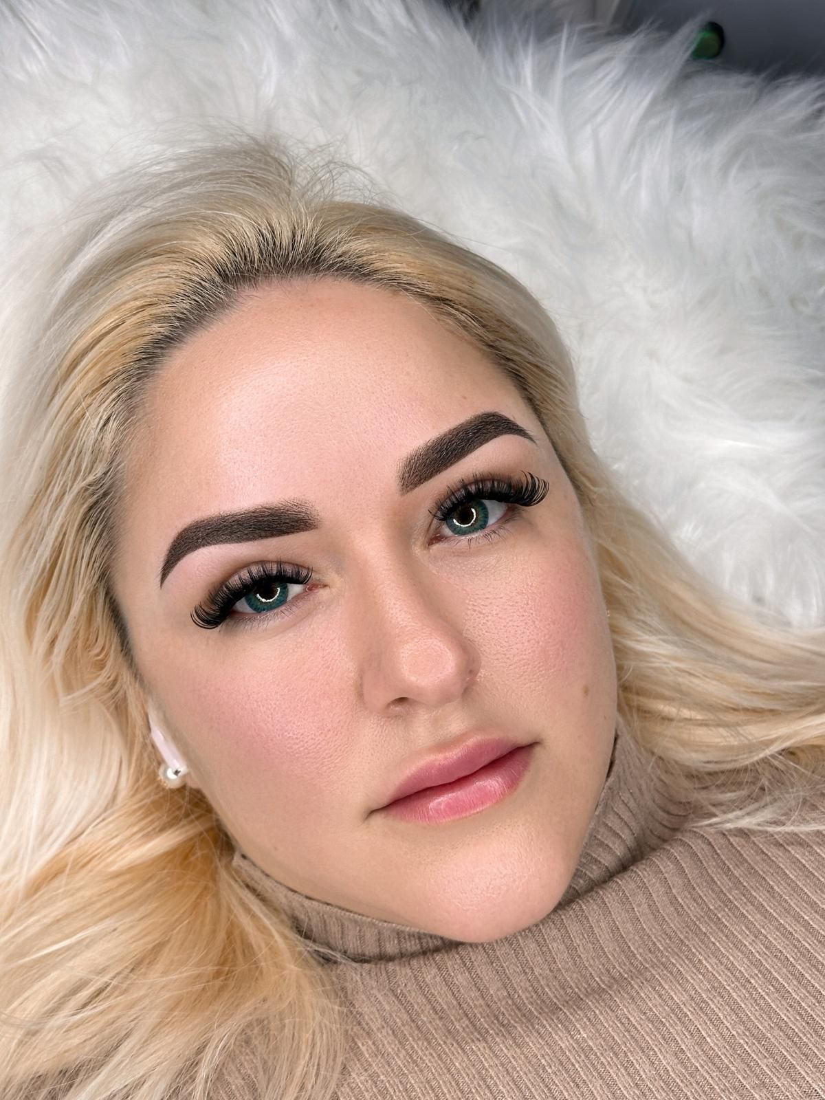 Woman with blonde hair, new eyebrow tattoo and false lashes, lying on white fur.