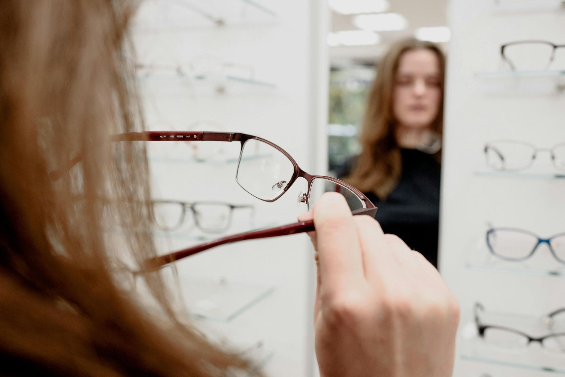 Hands trying on black eyeglasses in a display of assorted frames on a wall