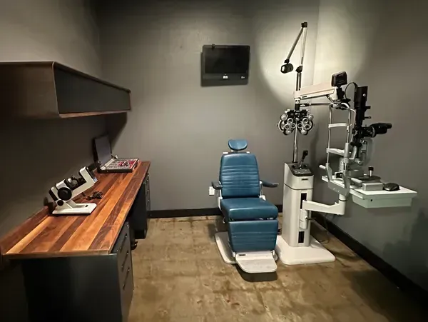 Optometry exam room with blue chair, eye chart, and diagnostic equipment on a counter.