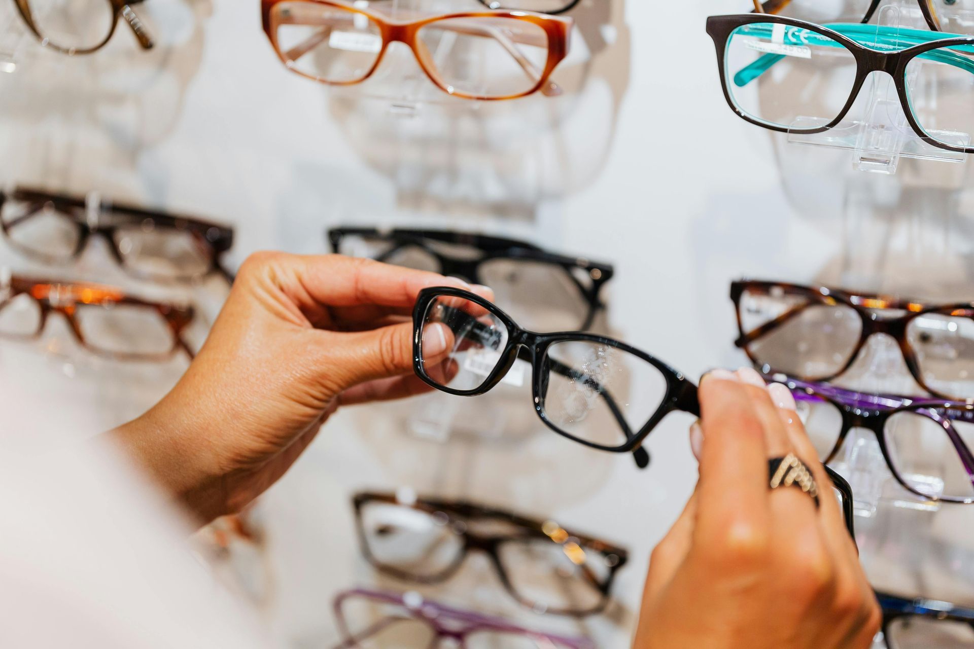 Hands trying on black eyeglasses in a display of assorted frames on a wall