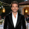 Hombre sonriente con traje de terciopelo negro en un elegante evento nocturno con guirnaldas de luces y velas.