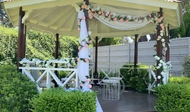 Cenador exterior decorado con sillas blancas, guirnaldas florales y una mesita en un entorno ajardinado.