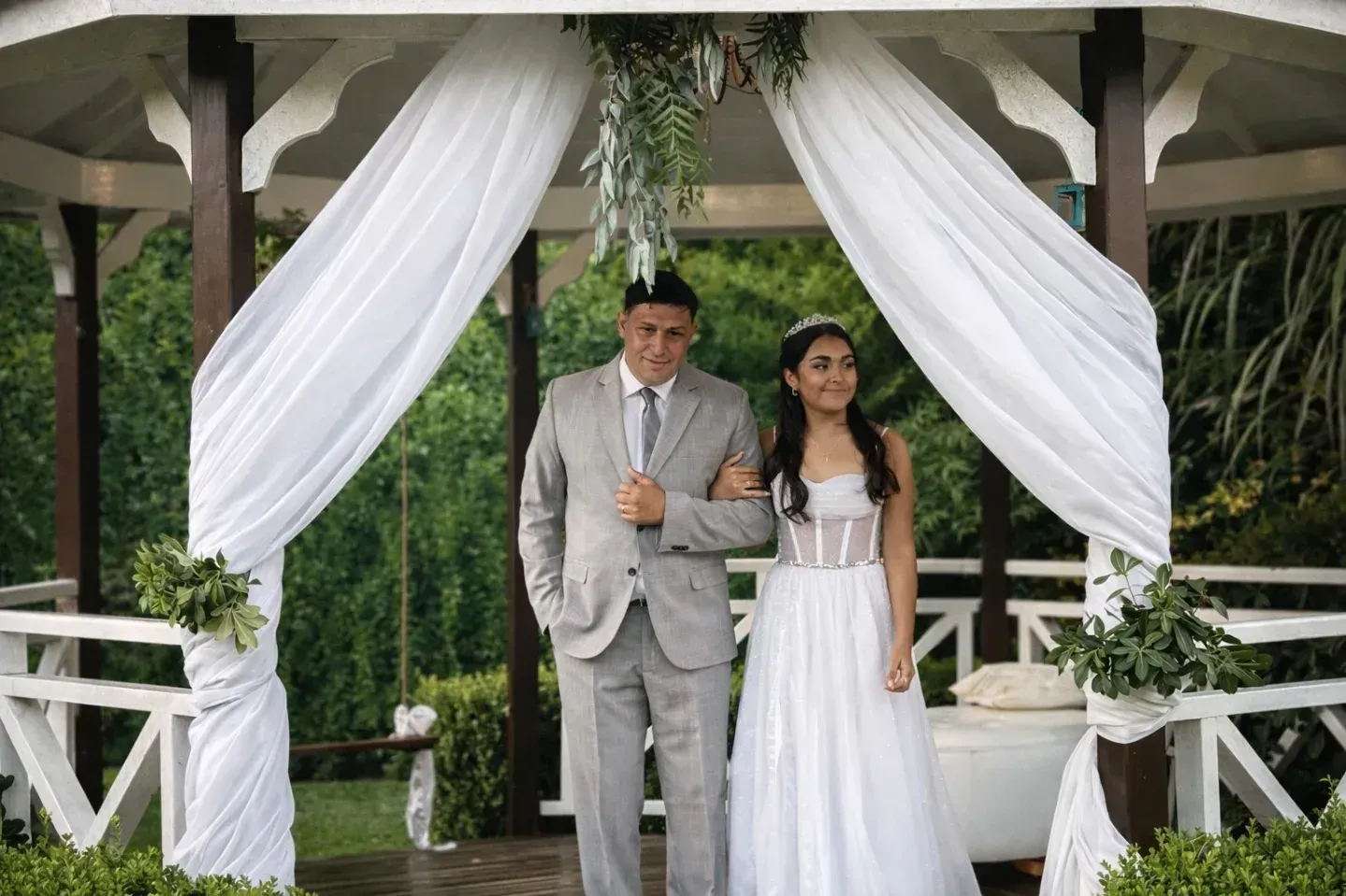 Novios de pie bajo una glorieta cubierta con tela blanca en un jardín.