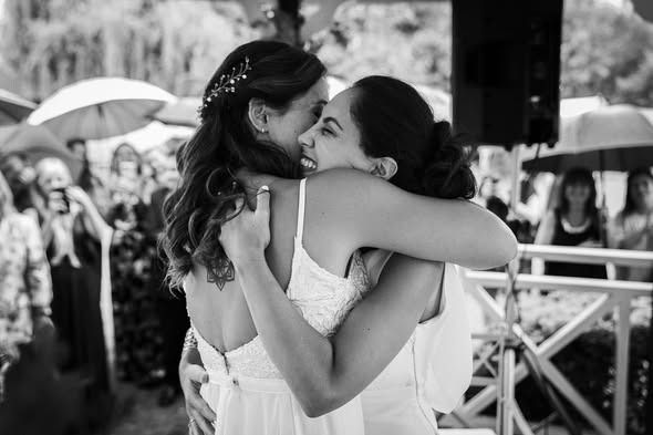 Dos personas se abrazan en una celebración al aire libre, con invitados y sombrillas al fondo.
