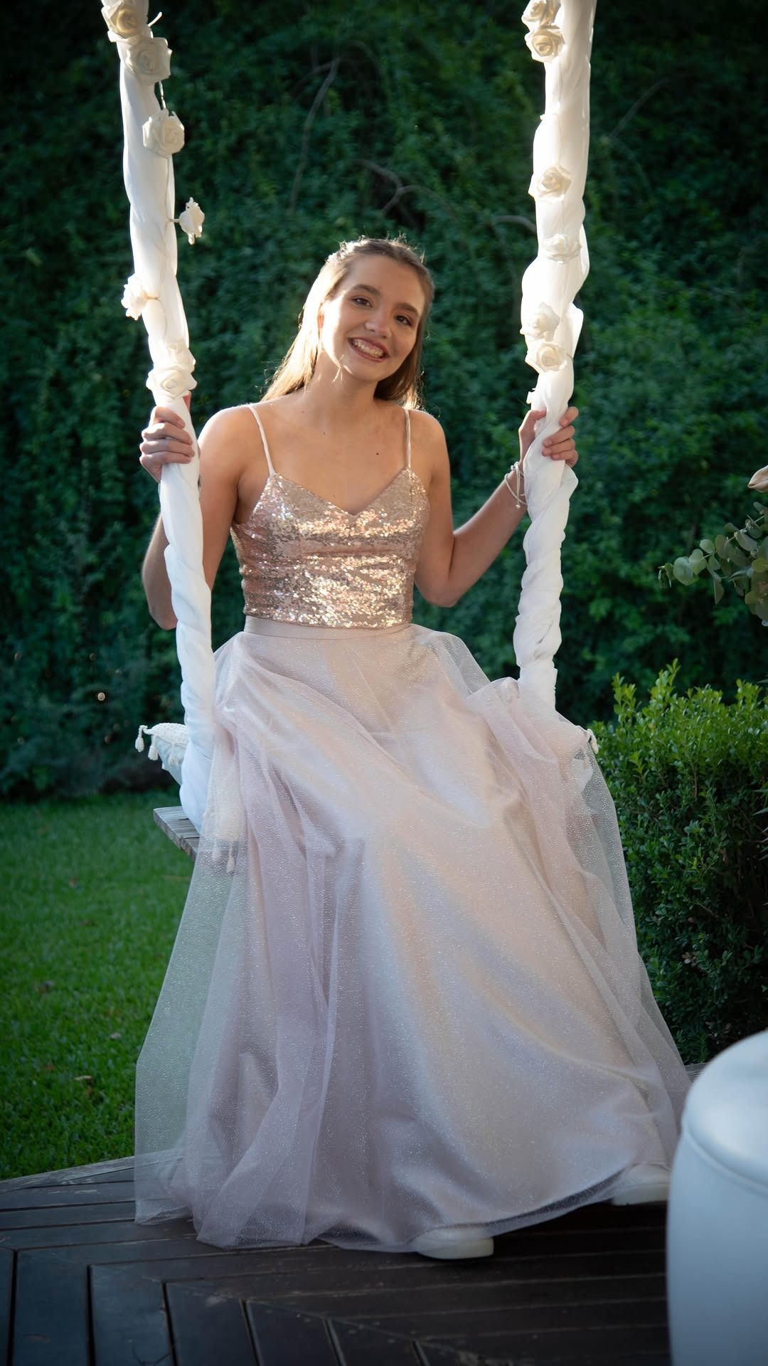 Mujer con un vestido dorado brillante sonriendo en un columpio decorado con flores en un jardín.