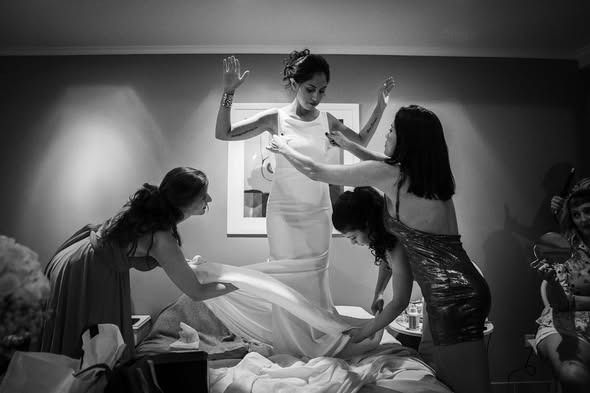 Cuatro personas colocan un vestido blanco sobre una mujer que está de pie sobre una cama en una habitación con poca luz.