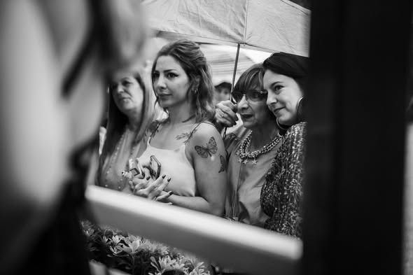 Tres mujeres de pie juntas bajo un paraguas, vistas a través del marco de una ventana en primer plano.
