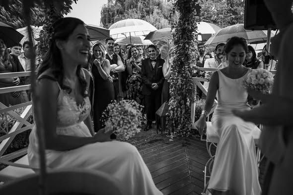 Dos mujeres con vestidos blancos sentadas en una ceremonia de boda al aire libre, sonriendo a los invitados cercanos.