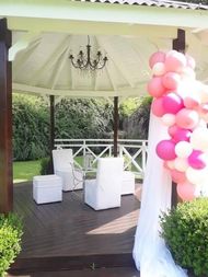 Cenador blanco con lámpara de araña, sillas blancas y arco de globos rosas en una terraza de madera al aire libre.