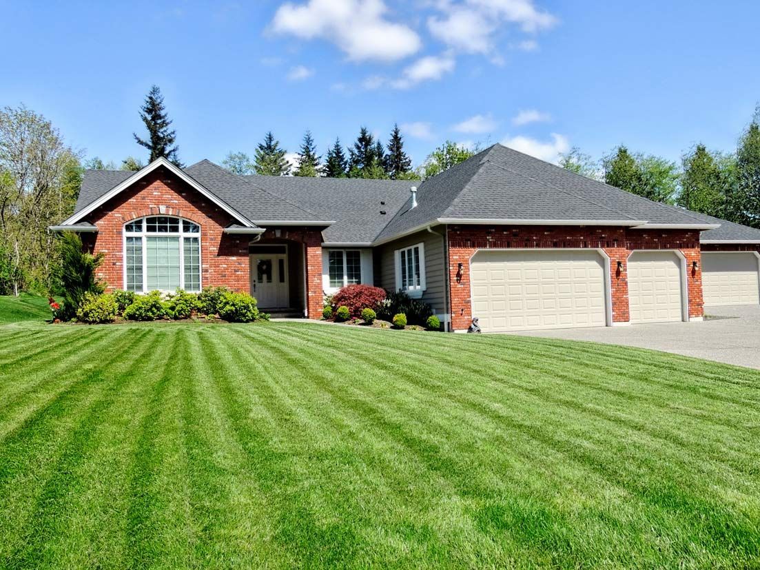 Well-manicured lawn with striped grass in front of a brick and gray roof house with a three-car garage. Blue sky.