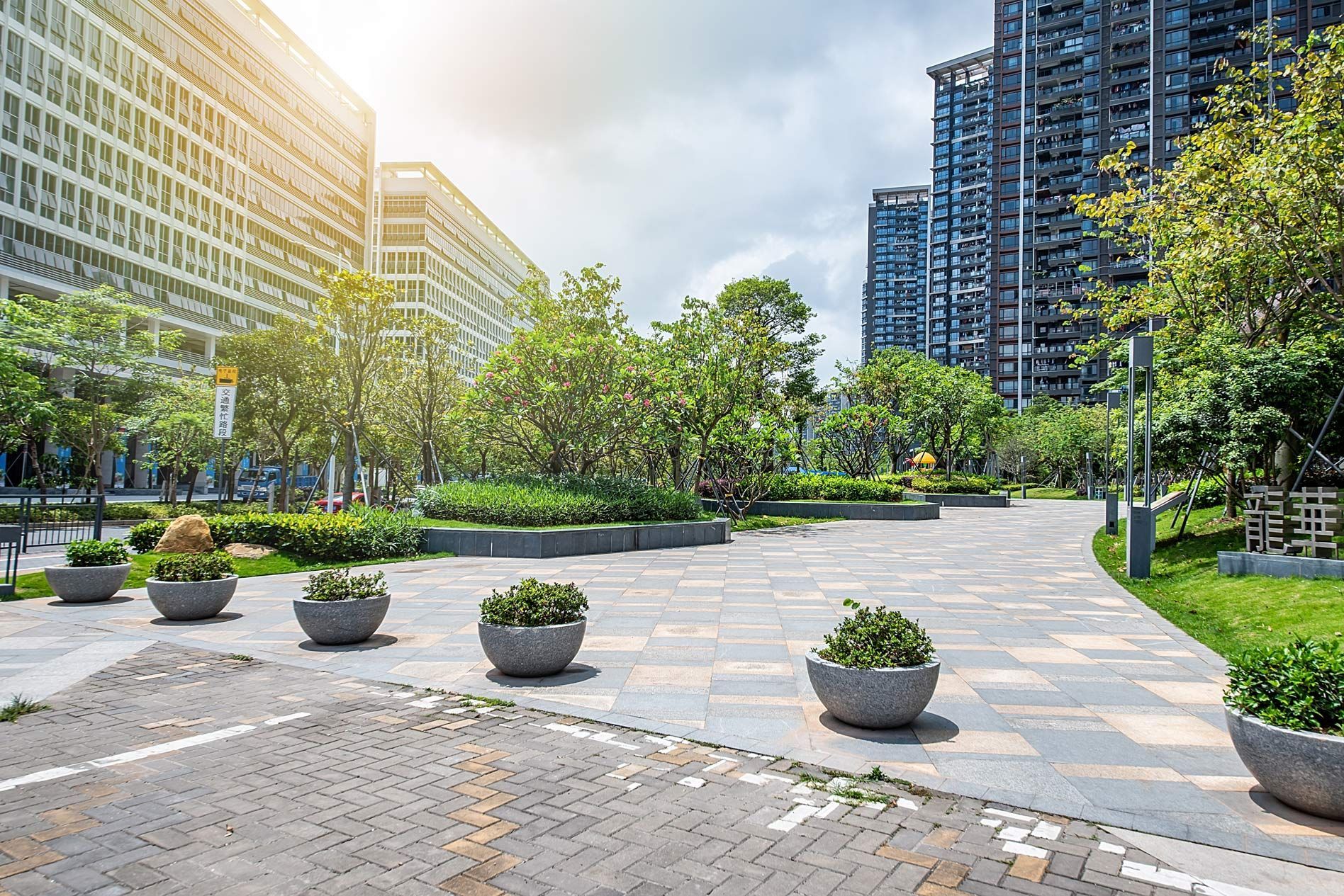 A paved urban plaza with large planters, green trees, and tall modern buildings.