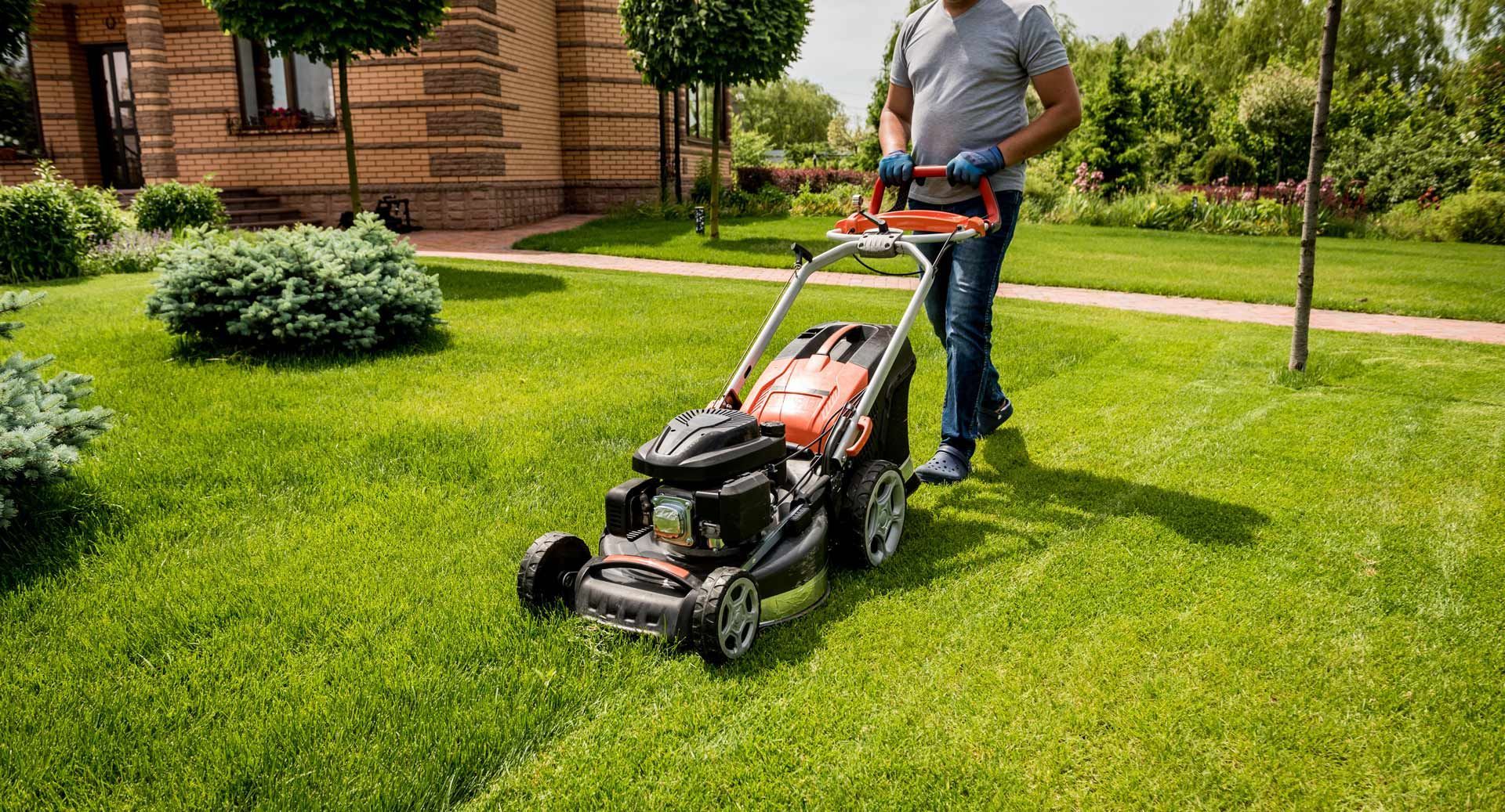 Man mowing a green lawn with an orange and black lawnmower in front of a house.