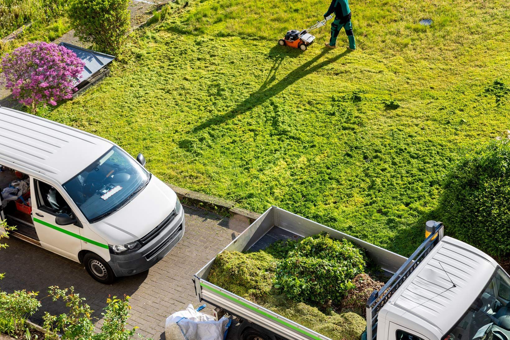 Lawn care crew: mowing grass with a mower, a truck with yard waste, and a van, on a sunny day.