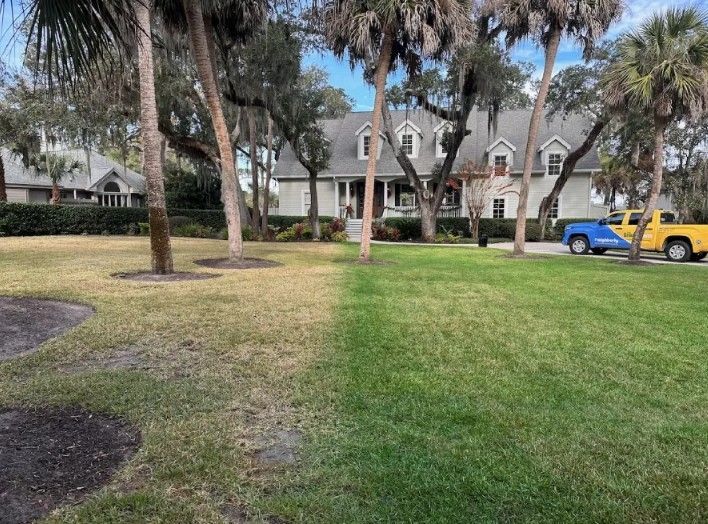 Lawn with patches of brown and green grass in front of a light-colored house. Two trucks are parked on the side. Lawn with patches of brown and green grass in front of a light-colored house. Two trucks are parked on the side.