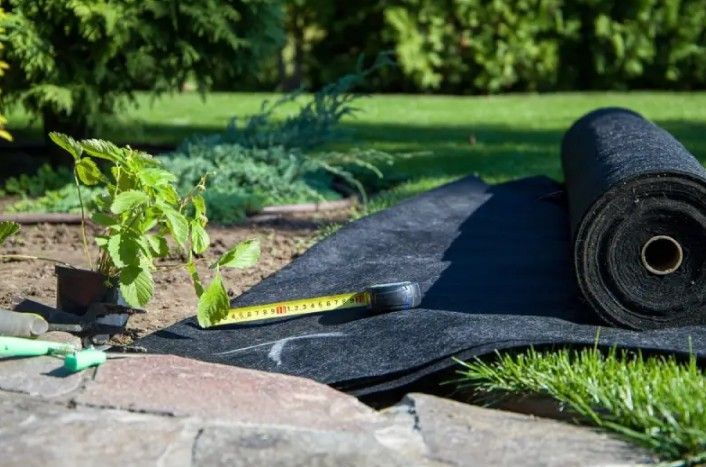 Black landscape fabric being installed in a garden bed with a measuring tape, near green plants. Black landscape fabric being installed in a garden bed with a measuring tape, near green plants.