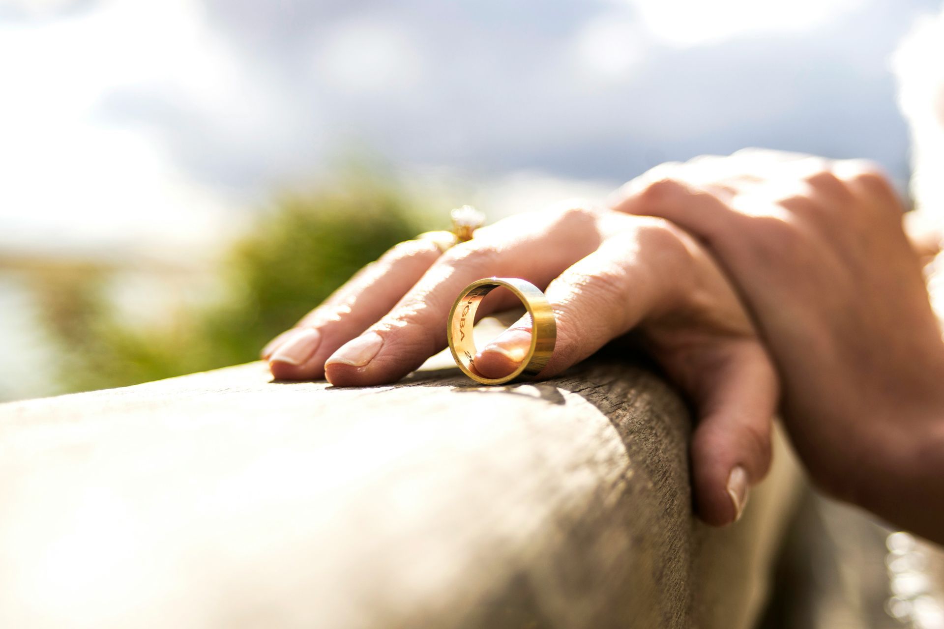 Hand with gold wedding ring on a wooden surface, blurred outdoor background.