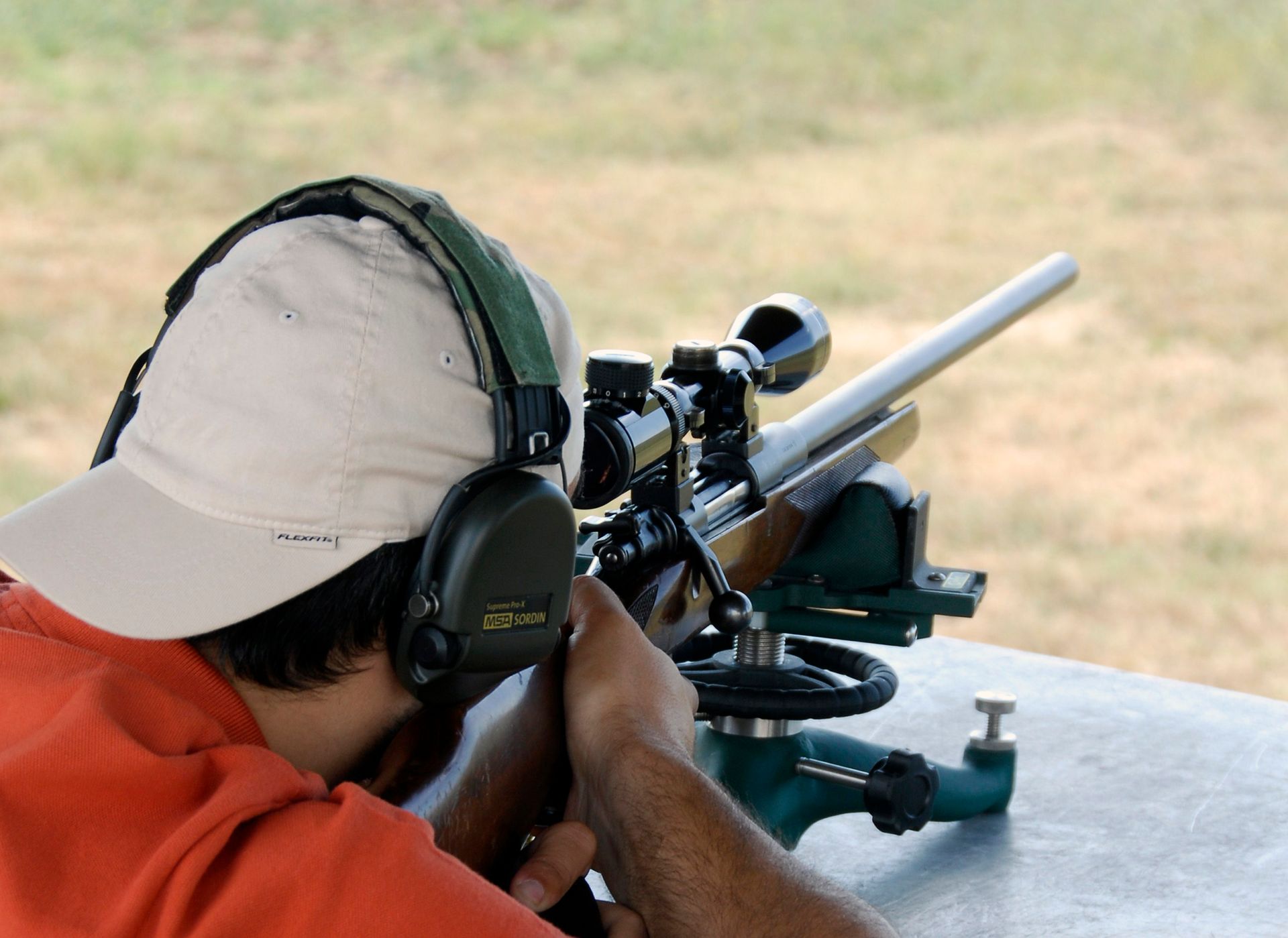 A person wearing a cap and ear protection aims a rifle mounted on a shooting rest at an outdoor range.