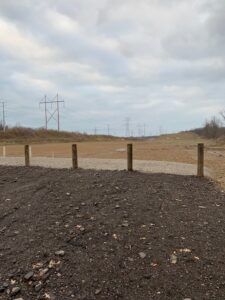 A row of wooden posts separates a dark gravel area from a flat, dry, open field under a cloudy, overcast sky.
