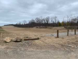 A wide, graded dirt field under a cloudy sky, with a line of wooden posts in the foreground and trees in the distance.