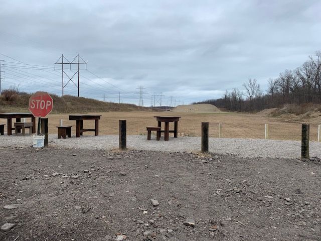 A gravel shooting range with wooden tables and a stop sign against a backdrop of hills and power lines under a cloudy sky.