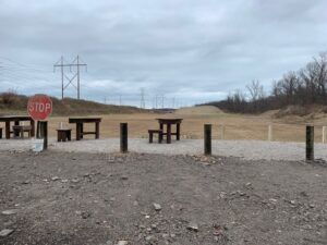 A gravel shooting range with several wooden tables, a stop sign, and a large dirt berm under a cloudy sky.