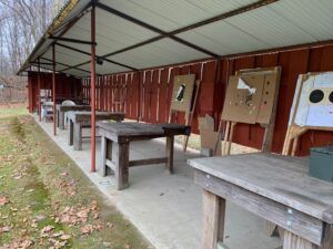 Outdoor shooting range with multiple wooden tables and target stands under a covered, red-walled structure.